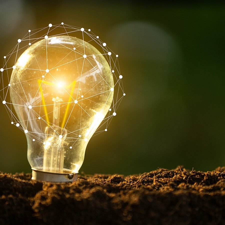 An illuminated light bulb on the ground with a geometric mesh overlay, set against a blurred outdoor background, symbolizing energy, innovation, and sustainable technology.