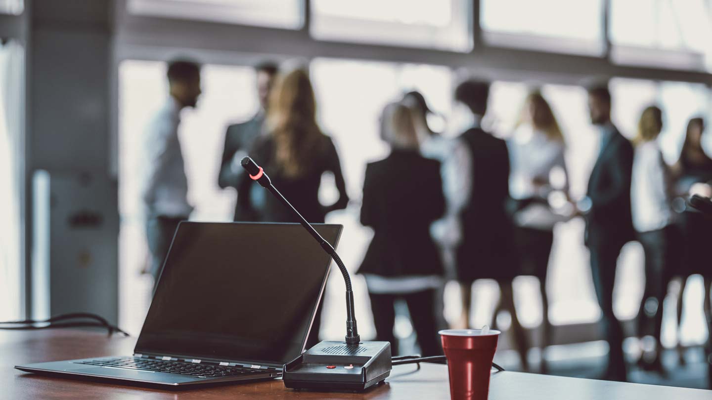 A laptop with an attached microphone on a table, set against a backdrop of blurred figures conversing, illustrating a professional gathering or meeting environment.