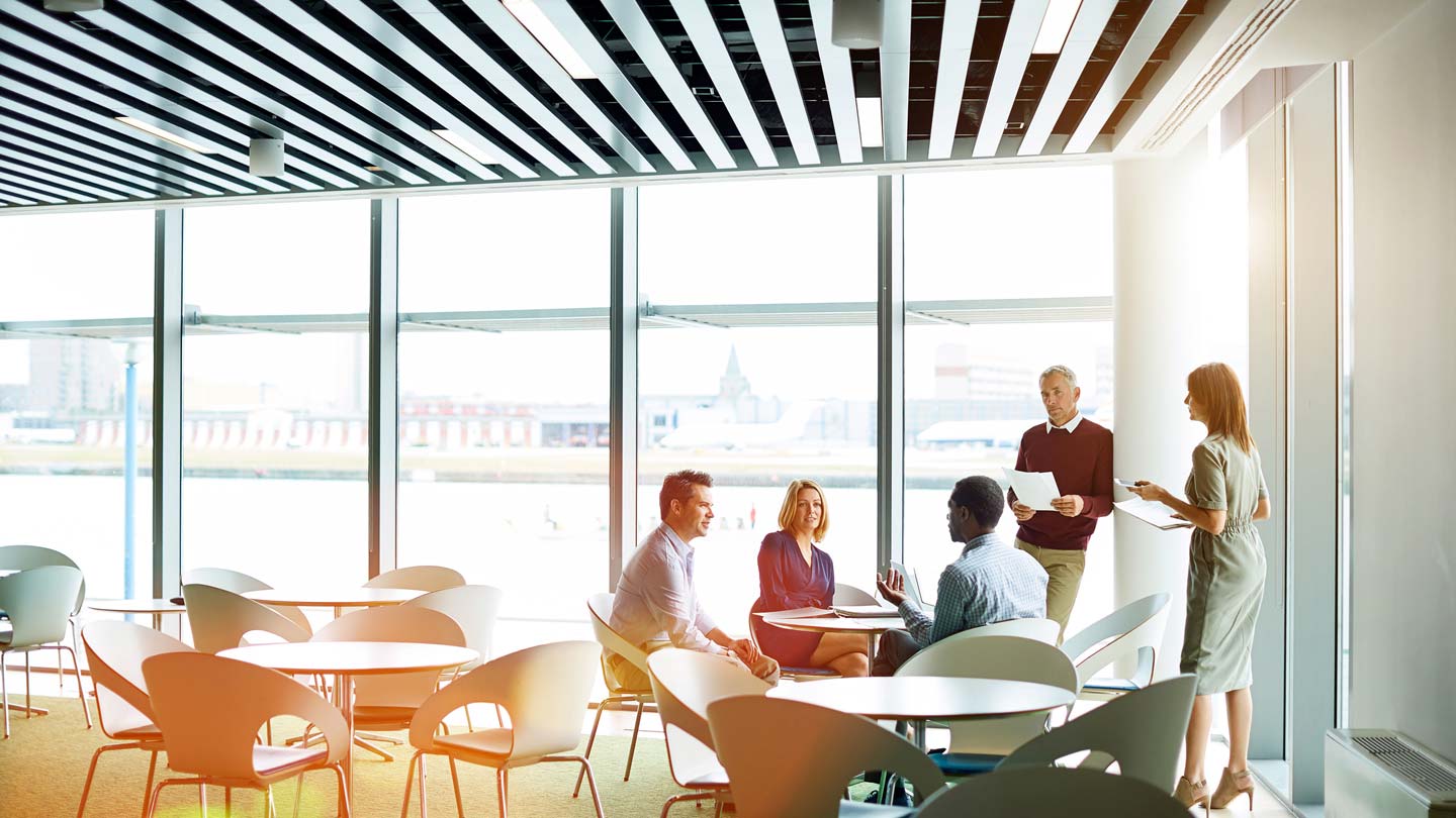 A modern office space with large windows, round tables, and a group of five individuals engaged in a discussion. The striking black and white striped ceiling contrasts with the warm sunlight, creating an inviting environment for collaborative work.