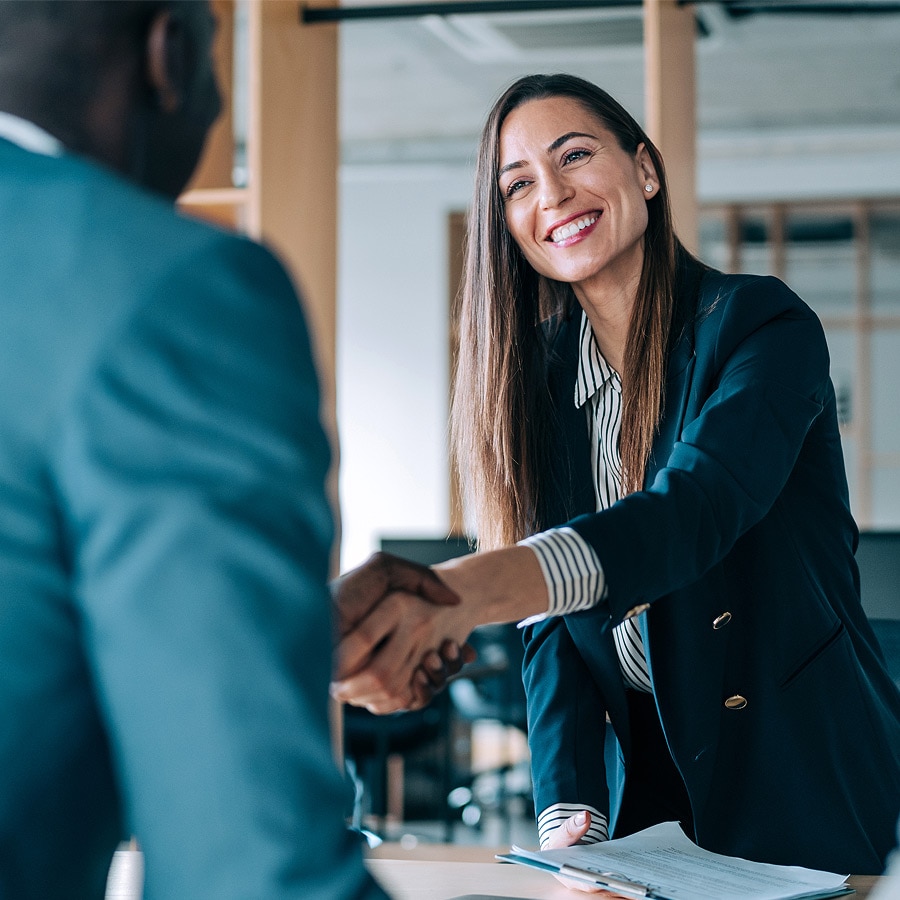 Two individuals shaking hands across a table in a professional setting, symbolizing agreement, introduction, or deal completion, with documents suggesting a business meeting.