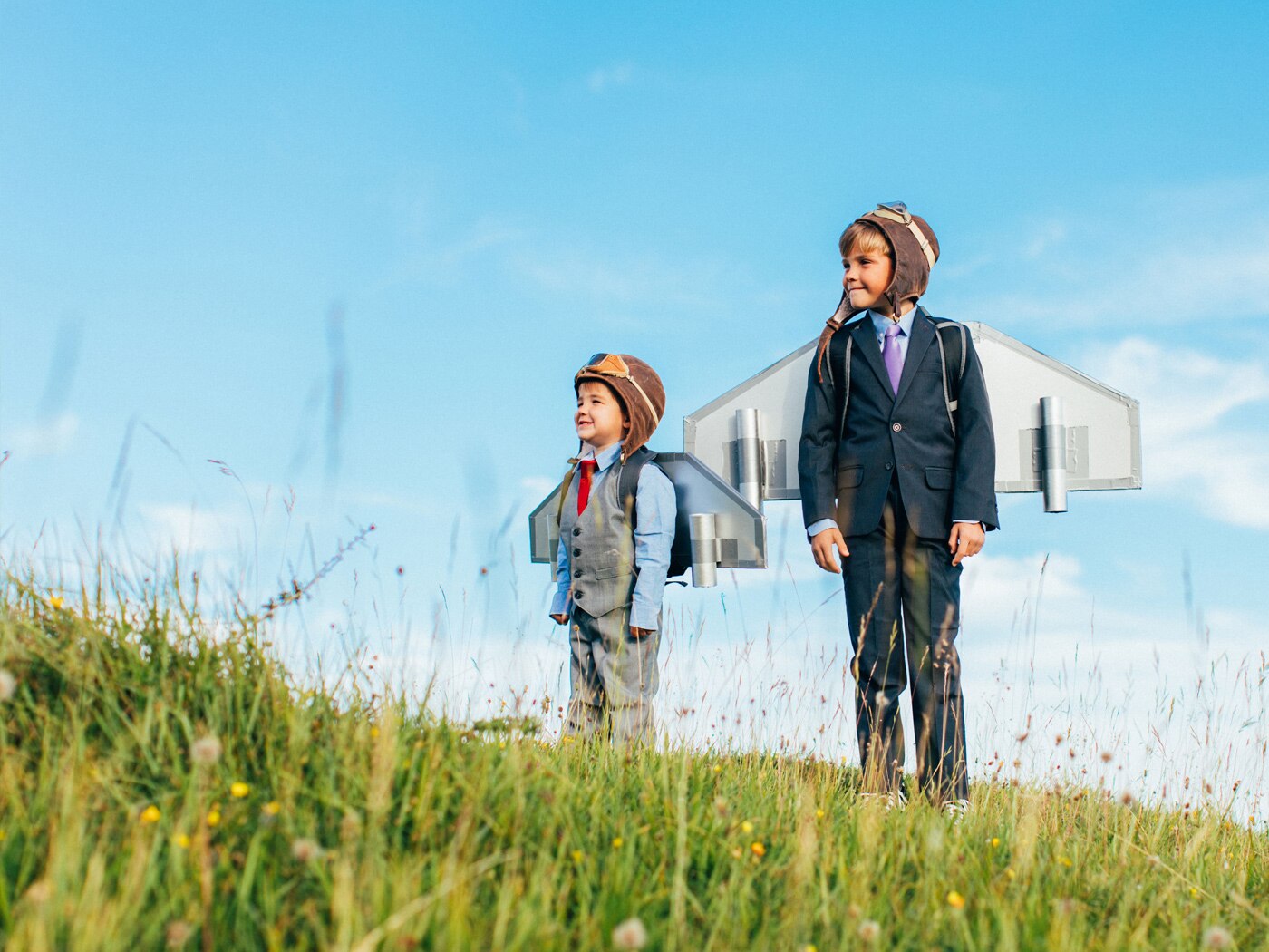 Two figures in a field under a clear blue sky, wearing suits and brown paper bags with drawn faces over their heads. The larger figure has wings attached, suggesting themes of anonymity, transformation, or freedom.