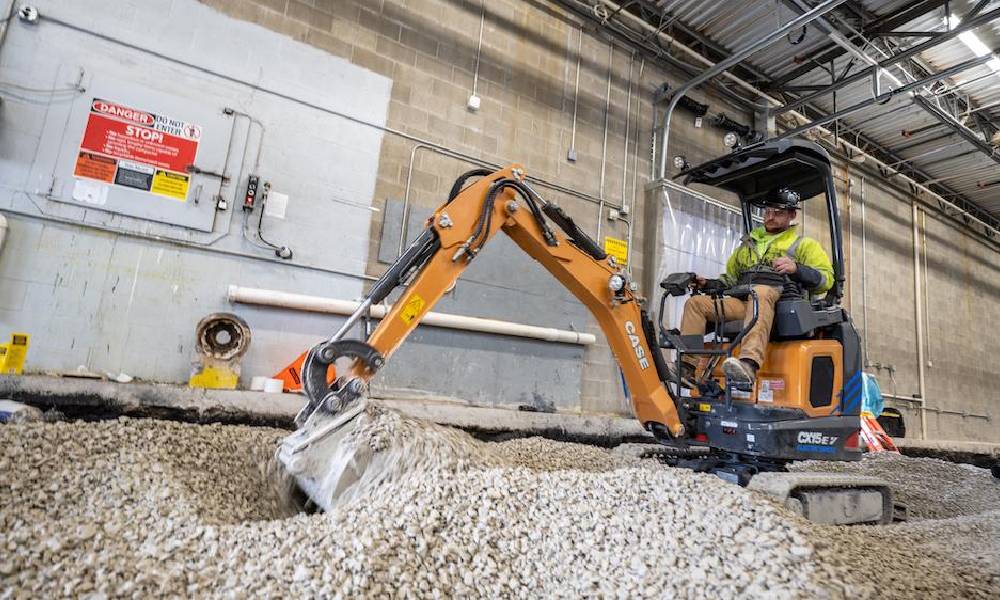 A small orange CASE excavator operated by a worker in a high-visibility vest and hard hat, moving gravel indoors in an industrial setting with concrete walls and a red fire alarm strobe light in the background.