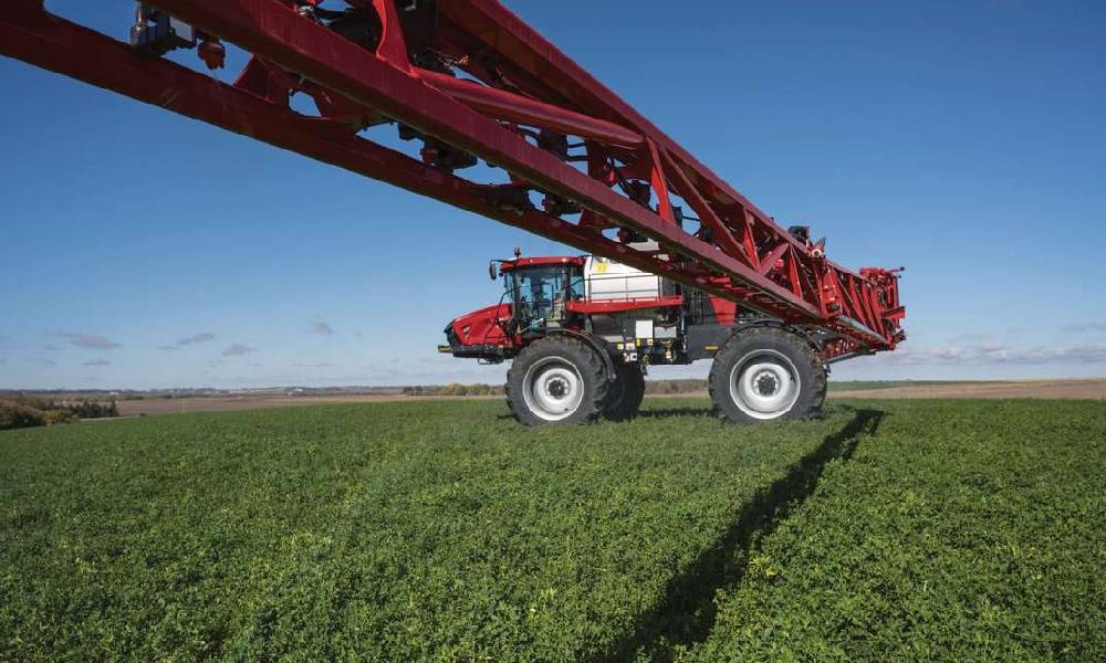 A large red Case IH agricultural sprayer with an extended boom operating in a green crop field under a clear blue sky. This image showcases modern farming technology used for efficient distribution of fertilizers or pesticides over large areas.
