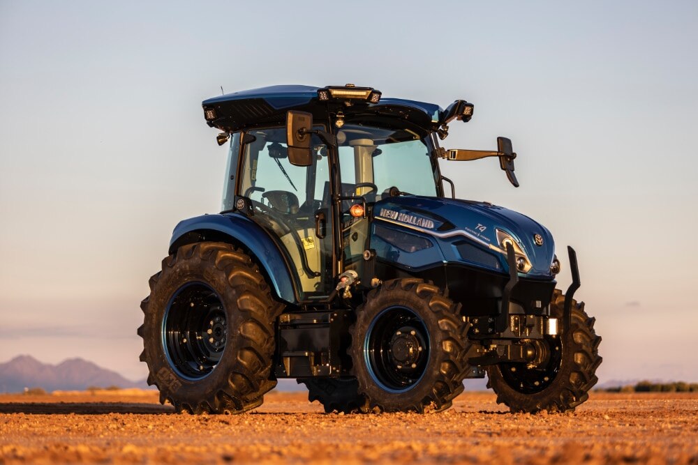 A modern blue New Holland tractor with large black tires, positioned in an open field during sunrise or sunset. The image highlights advanced agricultural machinery against a backdrop of a clear sky, showcasing modern farming practices.