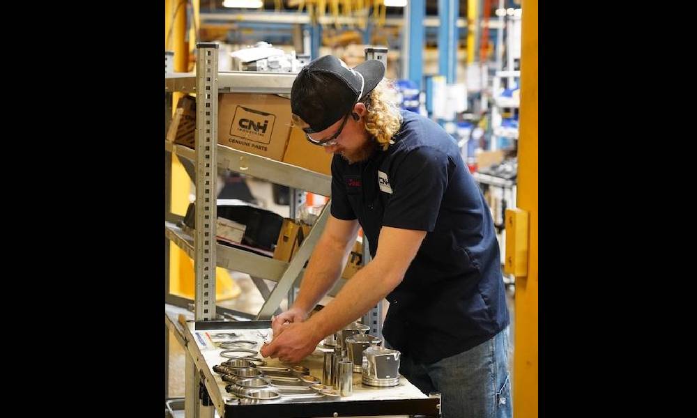 An individual working on a metal assembly line or workbench, surrounded by various metal parts and tools, in an industrial setting with yellow structures.